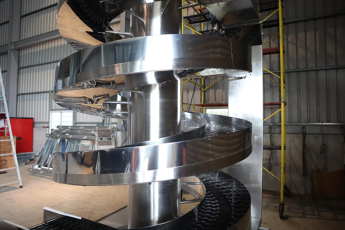 A stainless steel spiral conveyor with a black slat chain wound around a central column, standing in a workshop beside a mobile scaffold tower and stacked frame sections.