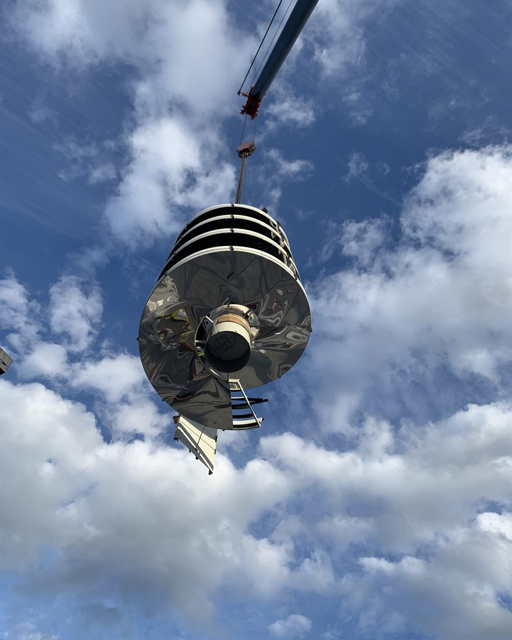 A stainless steel spiral conveyor section suspended from a crane against the sky.