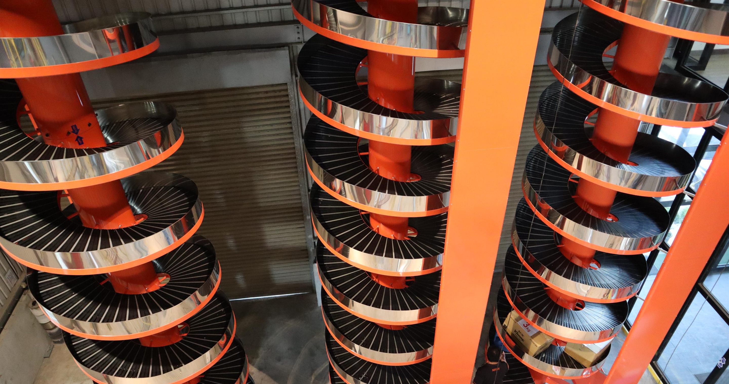 A row of NEXUS spiral conveyors standing in the factory before shipment, their stainless steel tiers wound around orange columns.