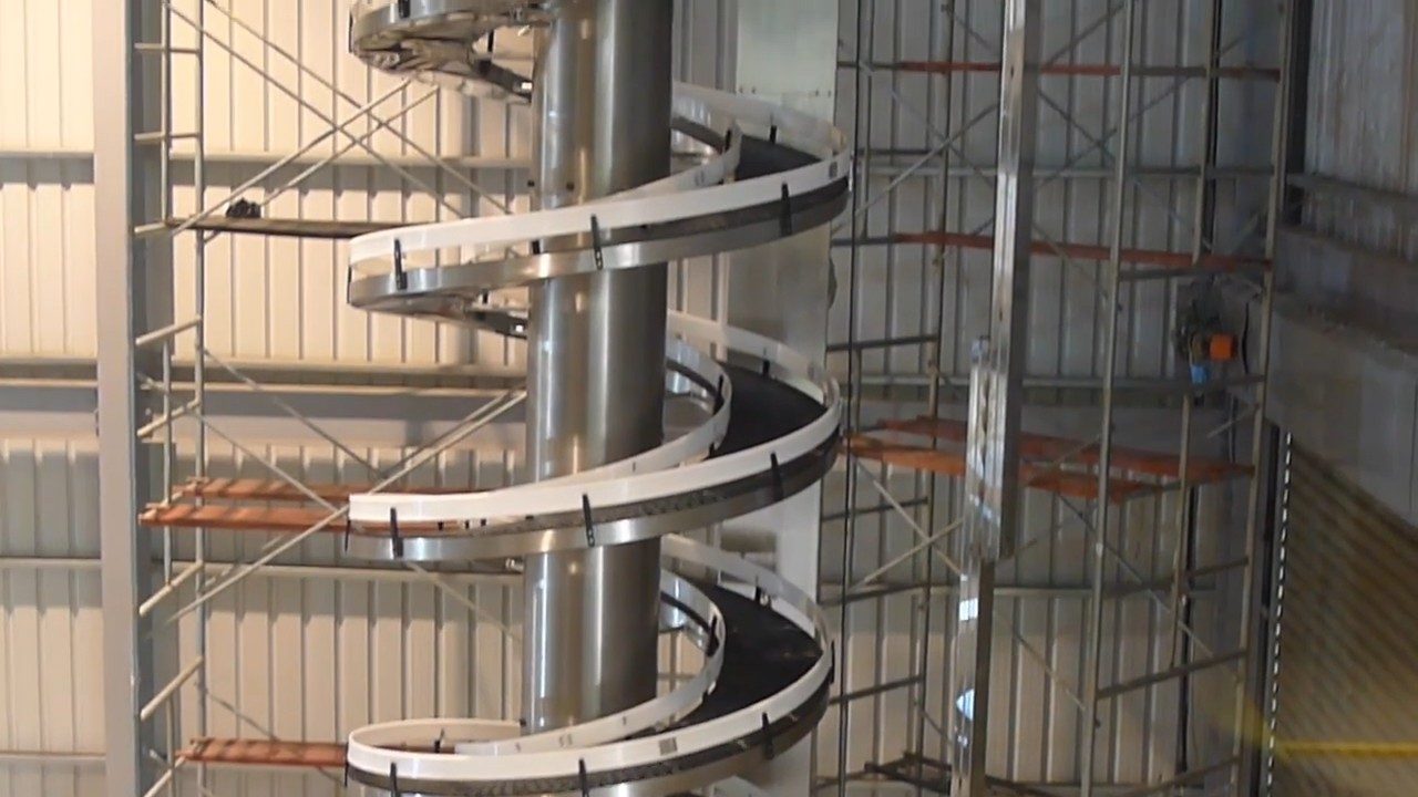 A stainless steel spiral conveyor with white side guards standing in a factory hall.