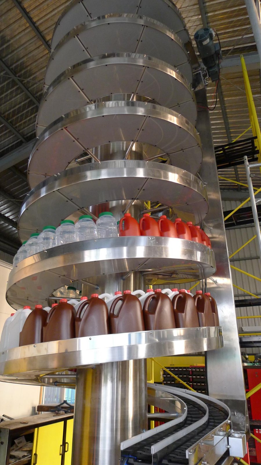 A dual track spiral conveyor seen from below, carrying rows of clear and brown plastic bottles on two separate tracks within one spiral structure.