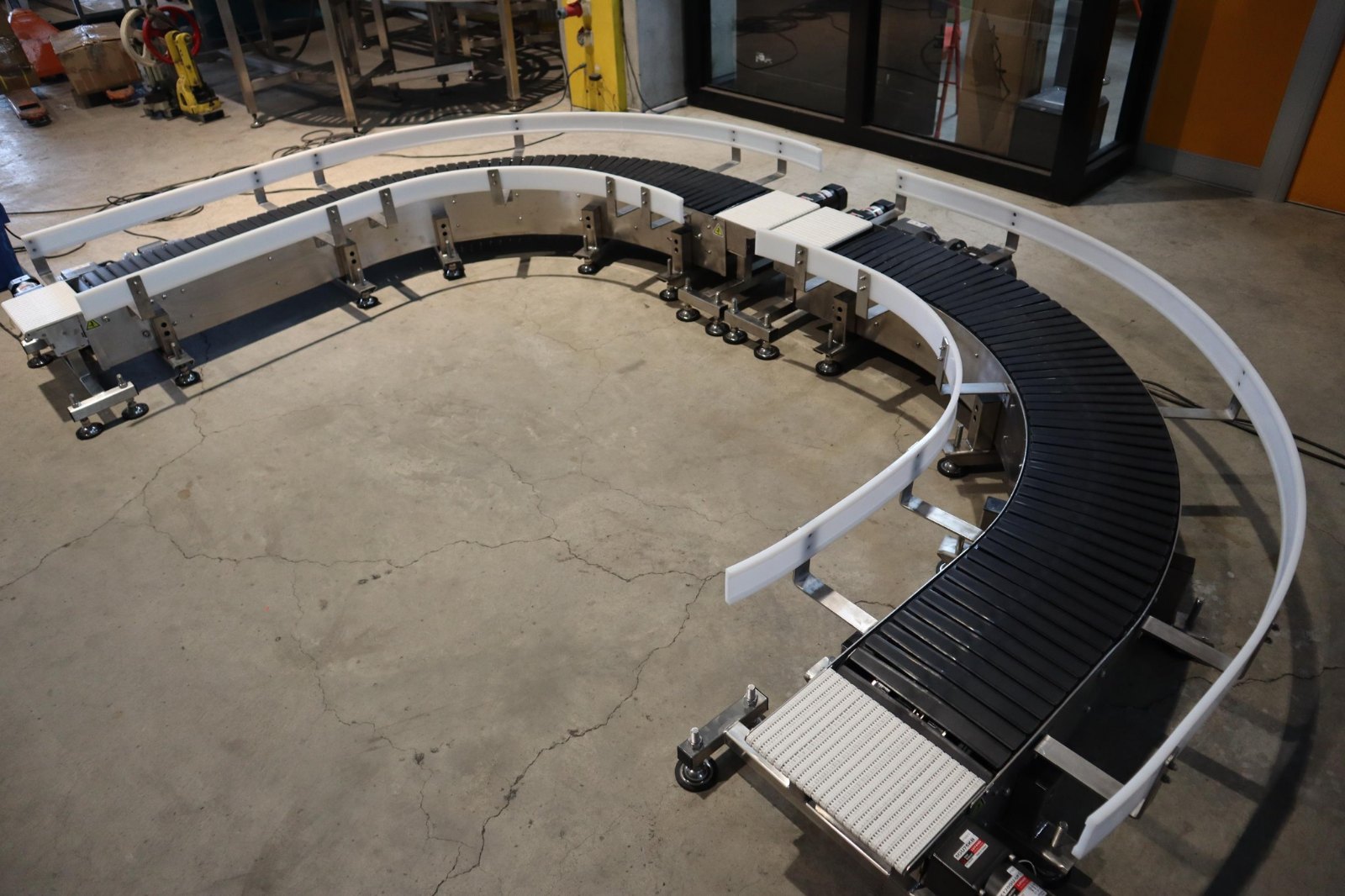 A side-flexing curve conveyor with a pale straight section joined to it at one end, standing on castors on the NEXUS factory floor.